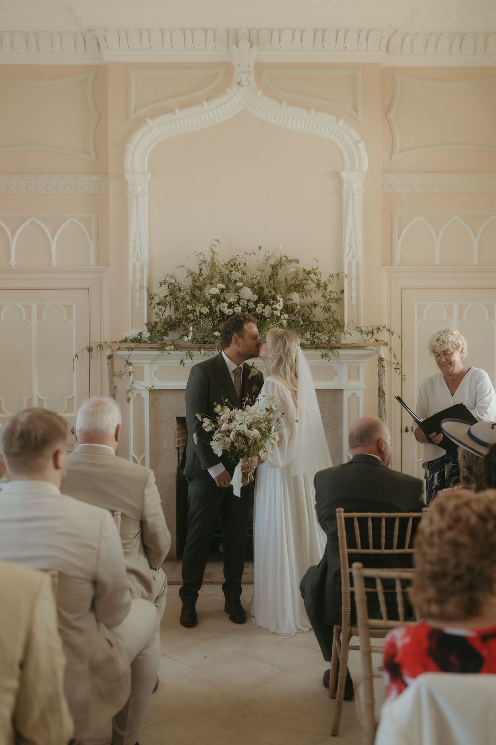 An elegant indoor wedding ceremony takes place in a white paneled room with ornate architectural details and floral decor.