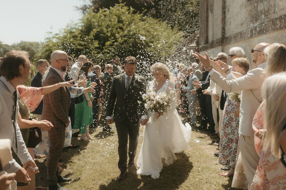 Wedding guests shower a couple with flower petals during a sunny outdoor ceremony exit along a garden path.