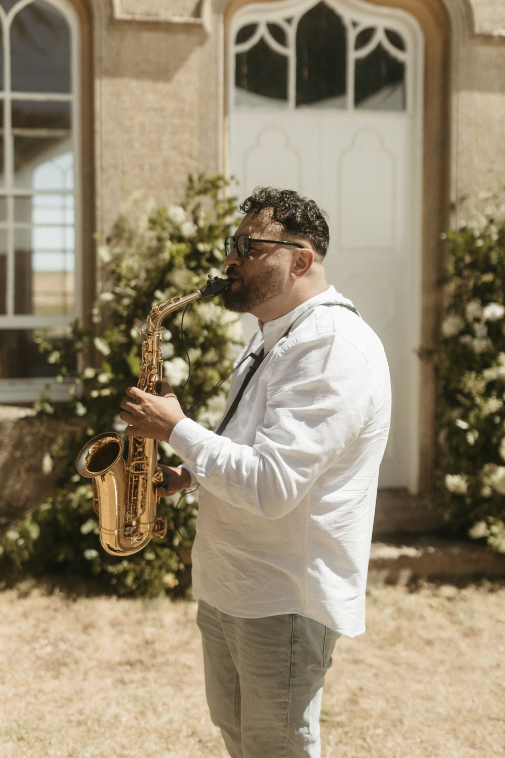 A musician playing a saxophone at an outdoor venue while wearing a white shirt and gray pants.