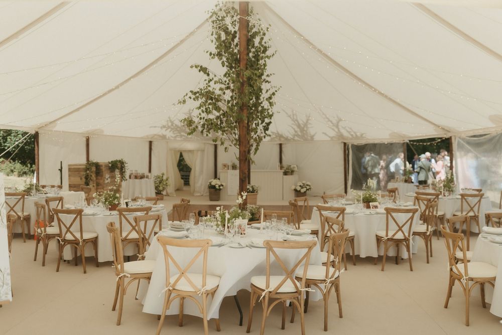 Elegant outdoor wedding reception setup with white tent, round tables, wooden cross-back chairs and rustic decor.