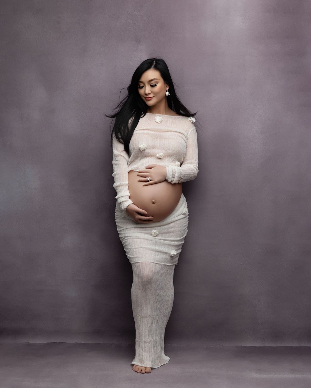 A maternity photo showing an expectant mother in a cream-colored dress standing against a gray backdrop.