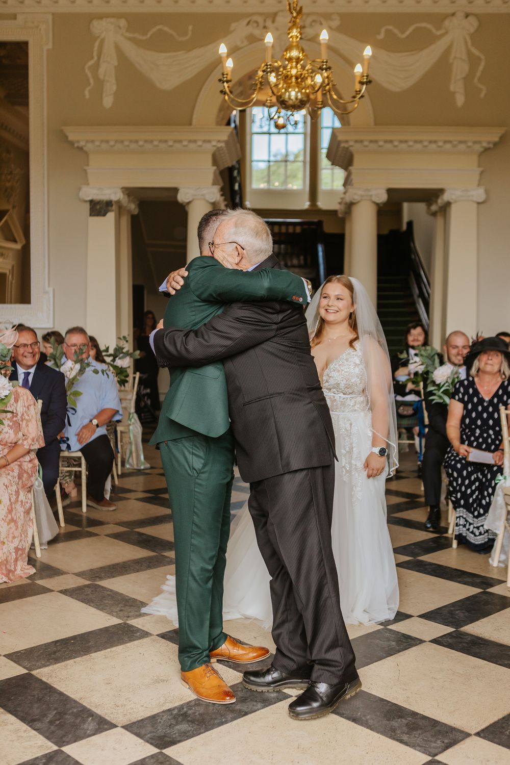 An emotional moment at an elegant wedding in a grand hall with checkerboard floors and a crystal chandelier.