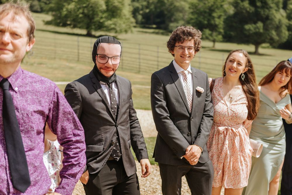 A wedding party in formal attire stands in a sunny field with green grass and trees in the background.