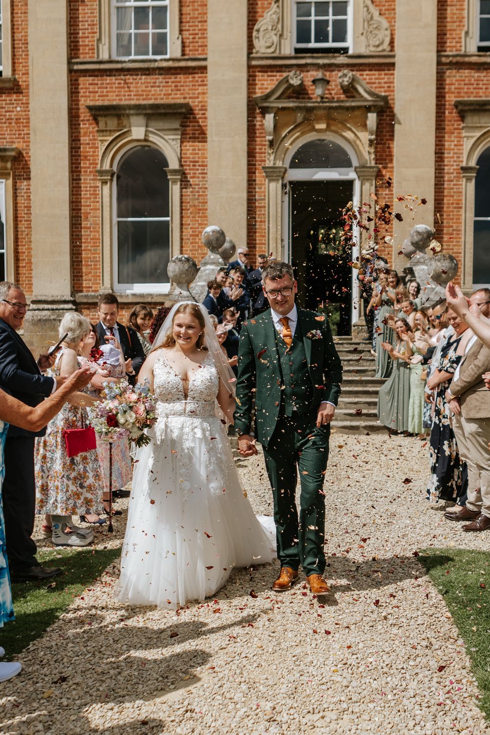 Wedding guests shower newlyweds with confetti outside a grand historic brick mansion with white columns.