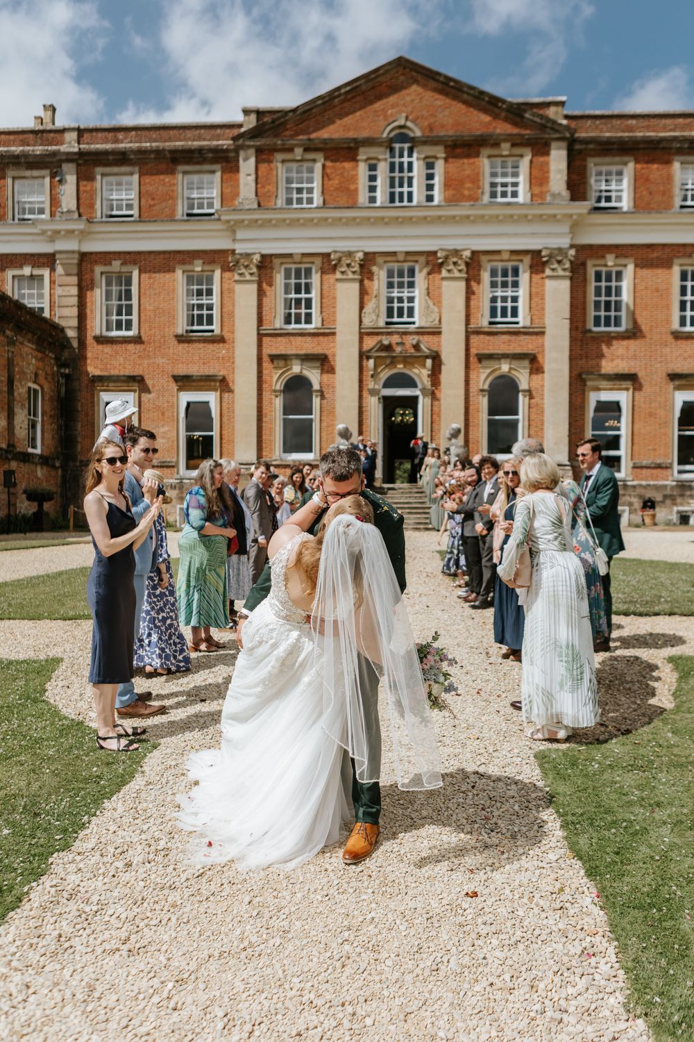 Wedding guests gather outside a grand red brick manor house during a summer celebration, creating a festive atmosphere.