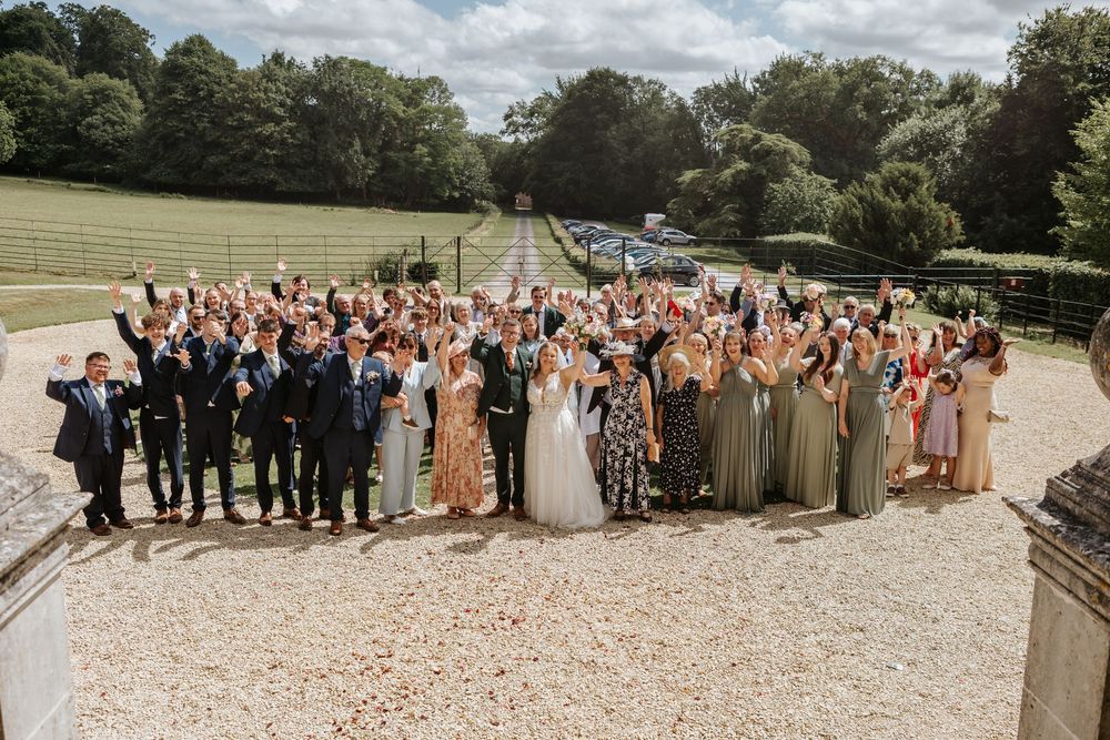 Large wedding party group poses outdoors on gravel path with trees and greenery in background.