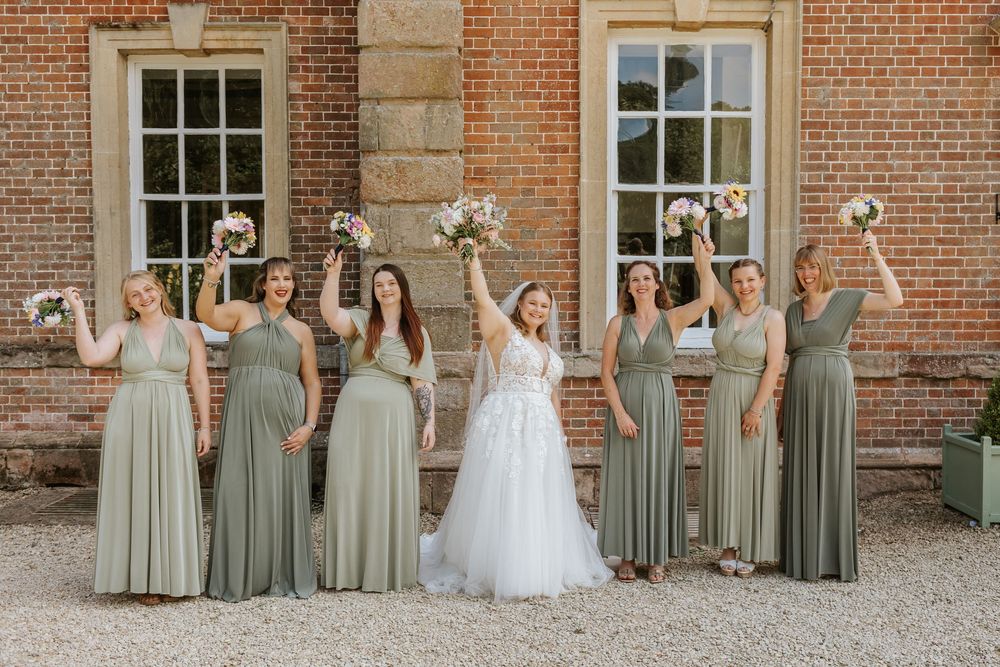 Wedding party stands in front of brick building with windows wearing sage green dresses and white gown.