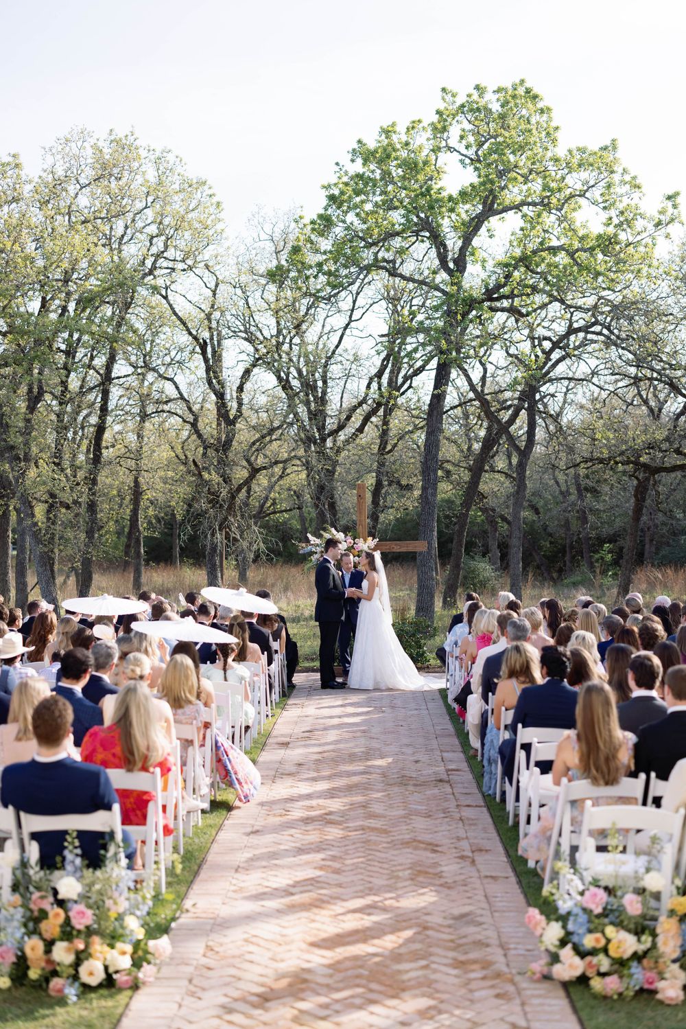 Outdoor wedding ceremony on a brick pathway surrounded by tall trees and white chairs decorated with floral arrangements.