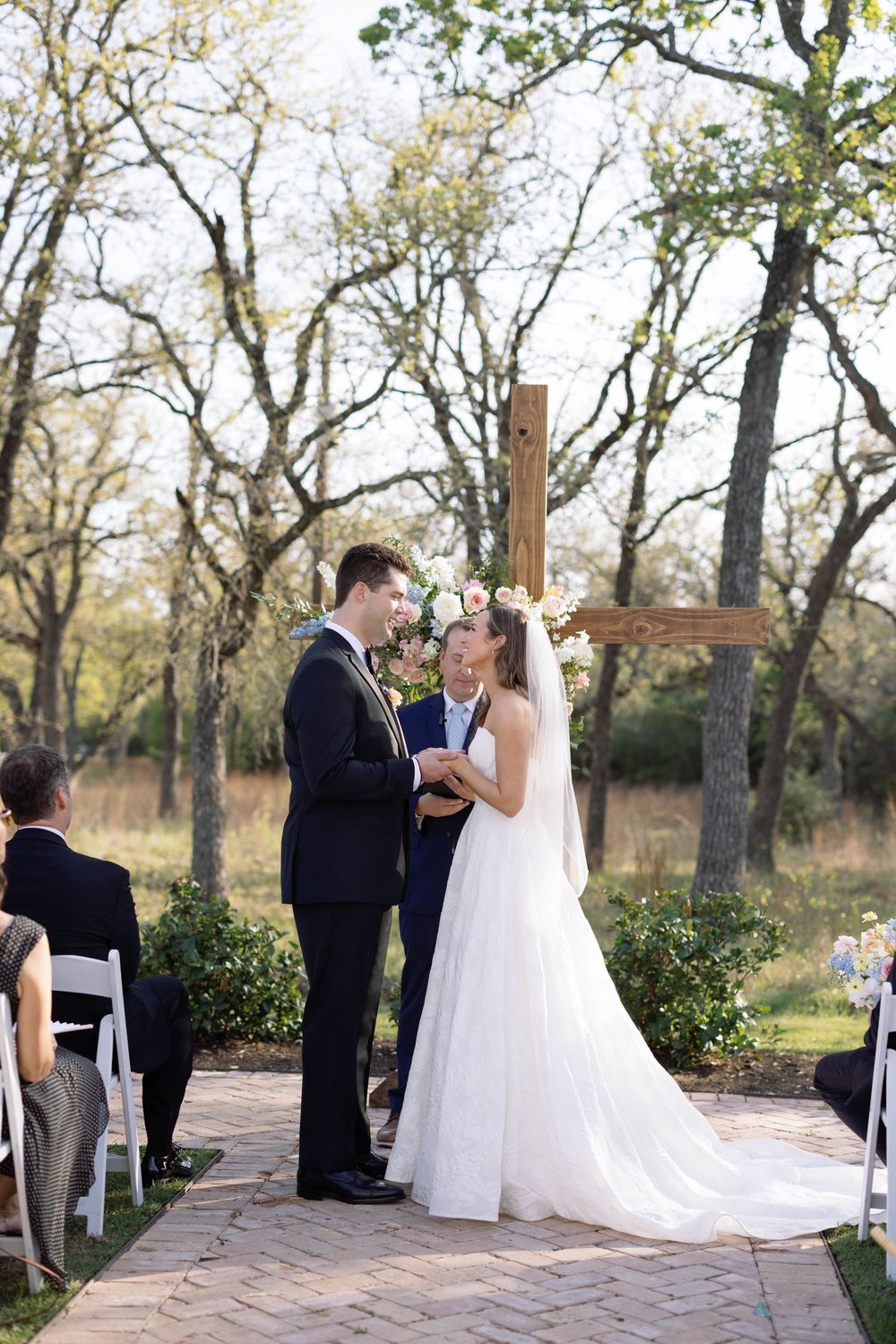 An outdoor wedding ceremony takes place on a brick path with a wooden cross and bare trees in the background.