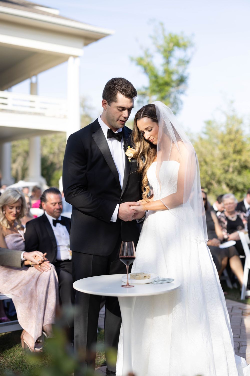 Bride and groom in formal attire share an intimate moment at their outdoor wedding ceremony near a table with wine glasses.