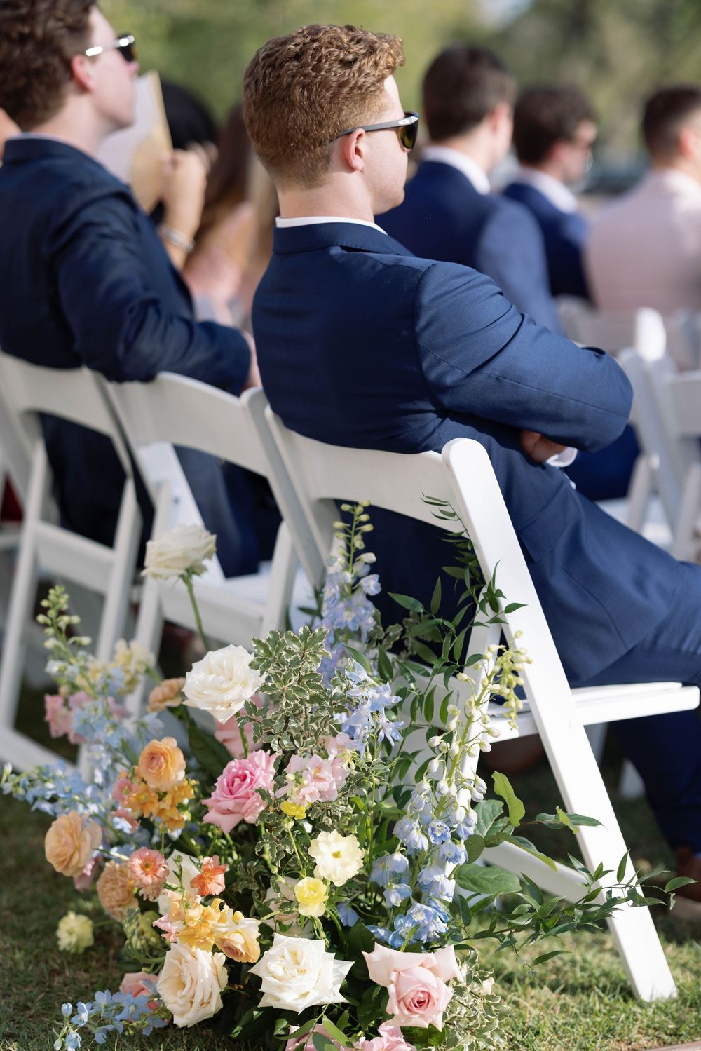 Peach and pink roses with blue delphinium flowers line the aisle at an outdoor wedding ceremony.