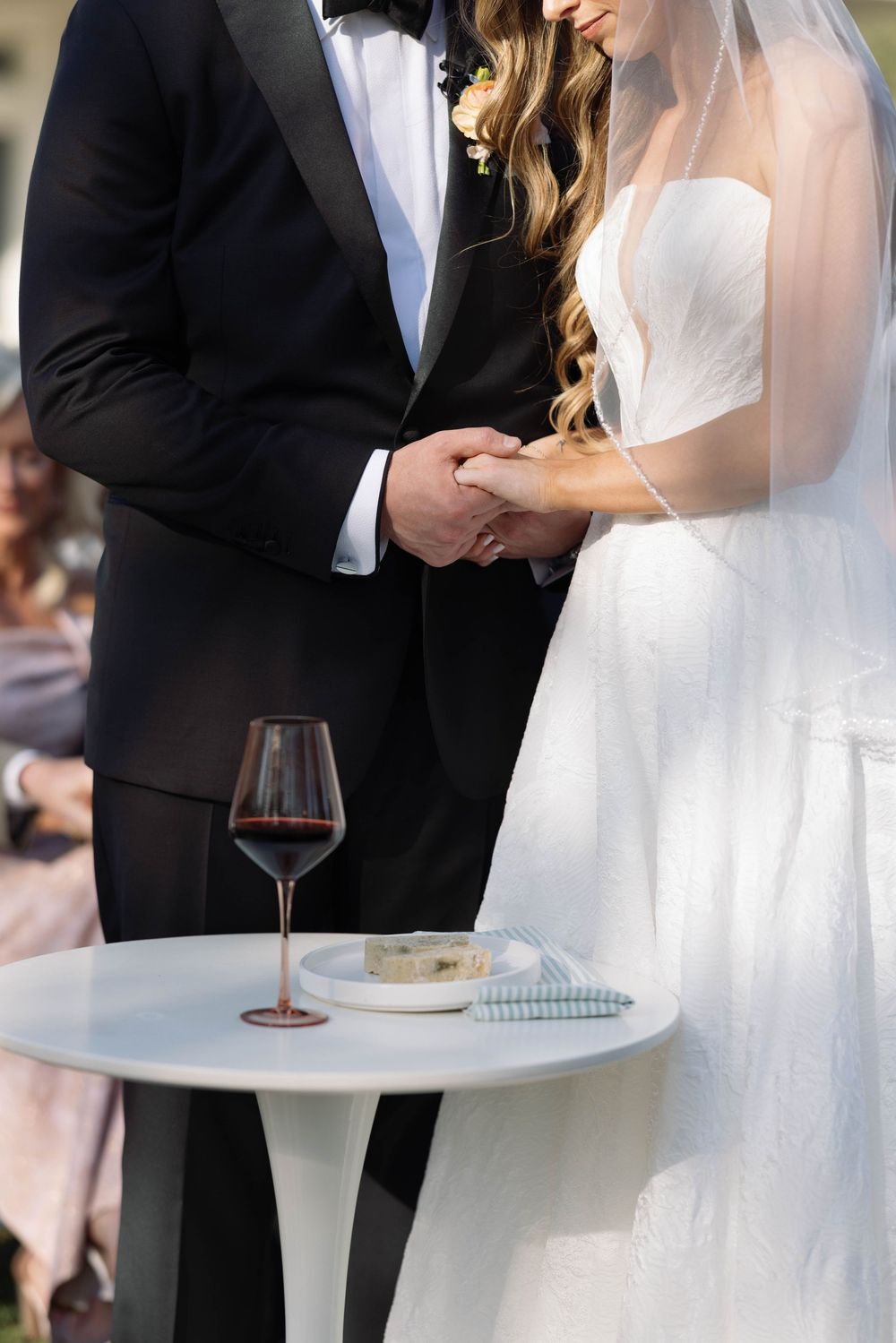 Close-up of newlyweds holding hands during ceremony with wine glass and small plate on white side table.