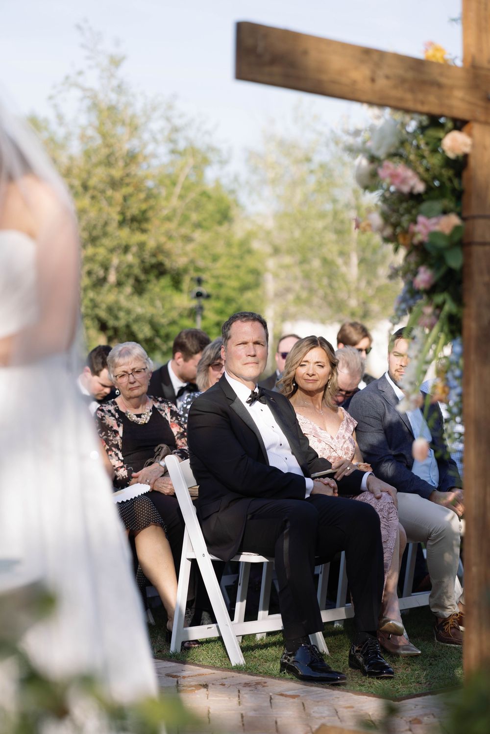 Wedding guests seated in white chairs during an outdoor ceremony under a wooden pergola structure.