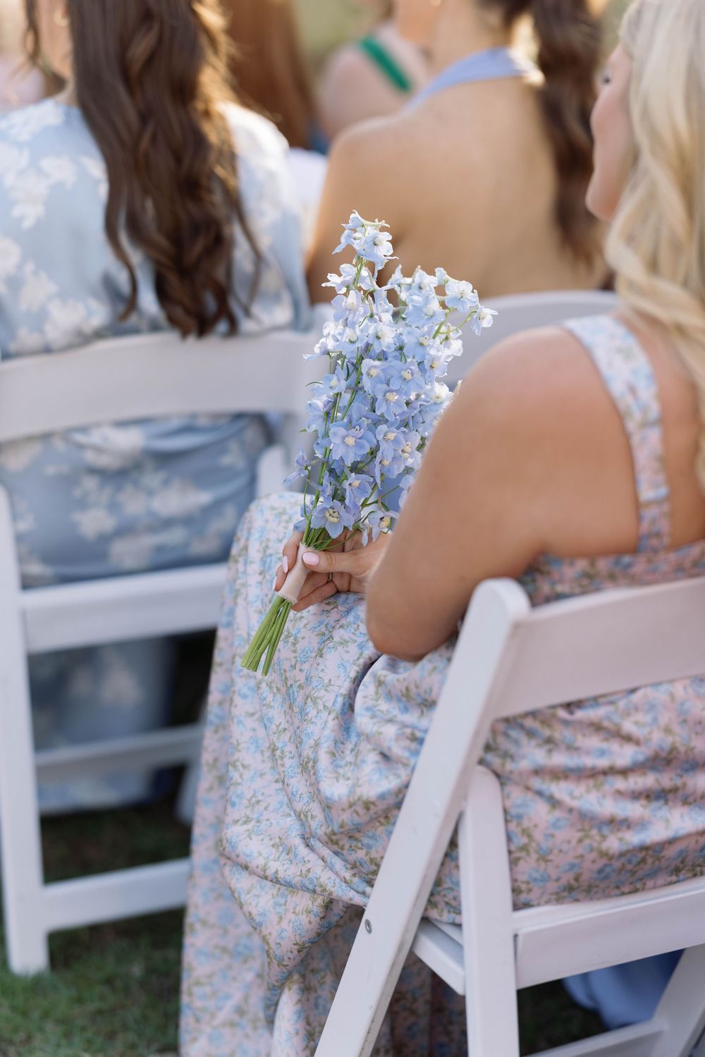 Person in a floral dress holds a delicate bouquet of light blue delphinium flowers while seated in a white folding chair.