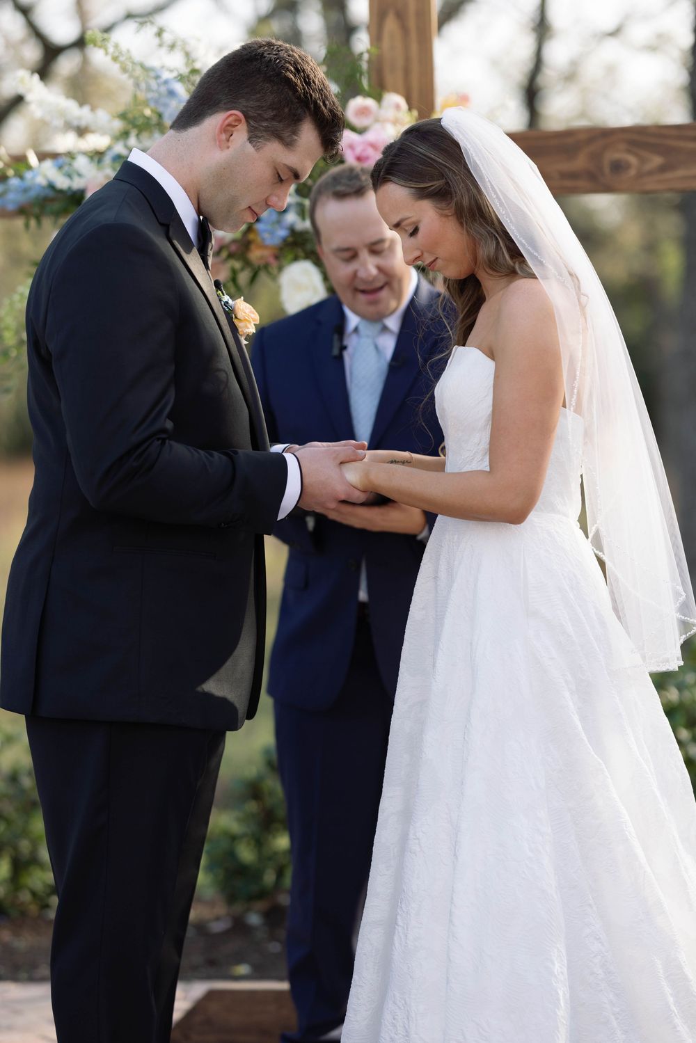 A wedding ceremony takes place outdoors with a bride in white dress and groom in black suit exchanging rings.