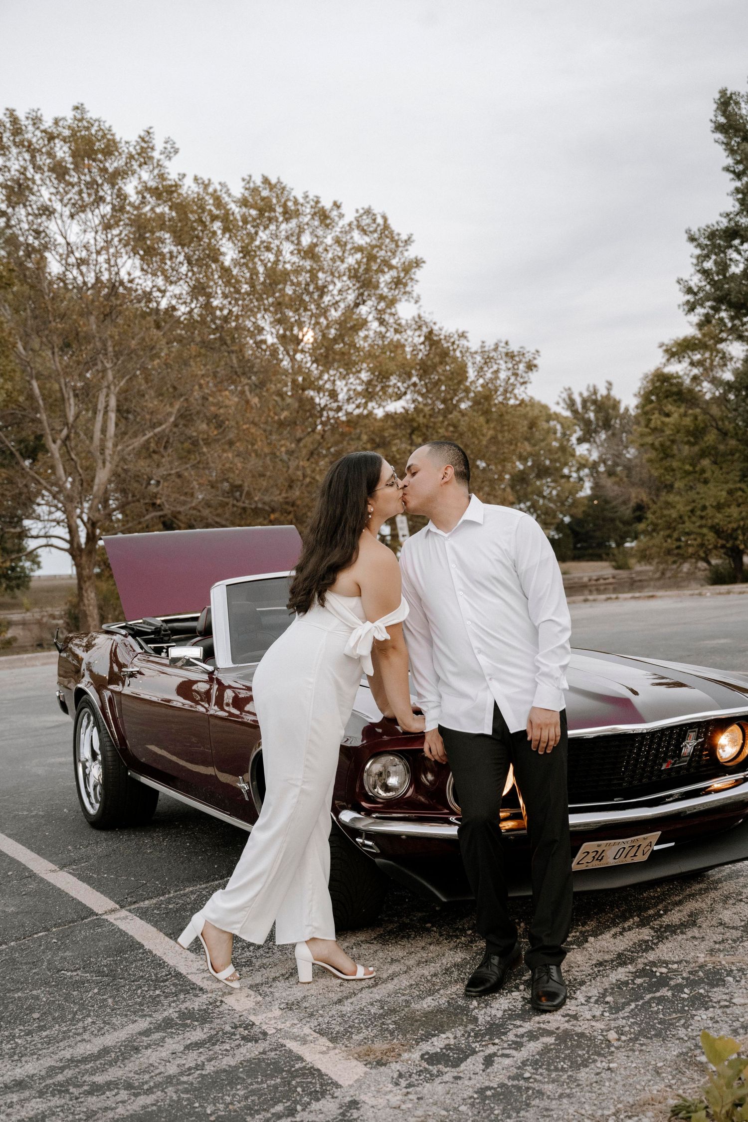 A romantic couple shares a kiss while leaning against a vintage burgundy Ford Mustang on a cloudy autumn day.