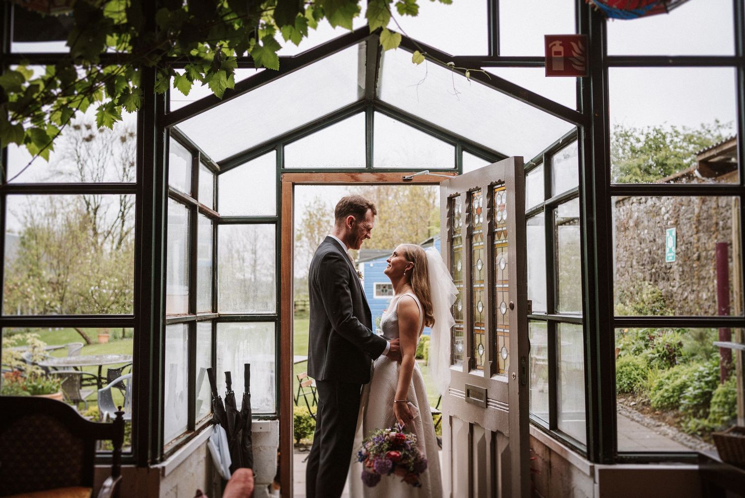 A romantic couple stands embracing in a glass greenhouse doorway during a garden wedding ceremony.