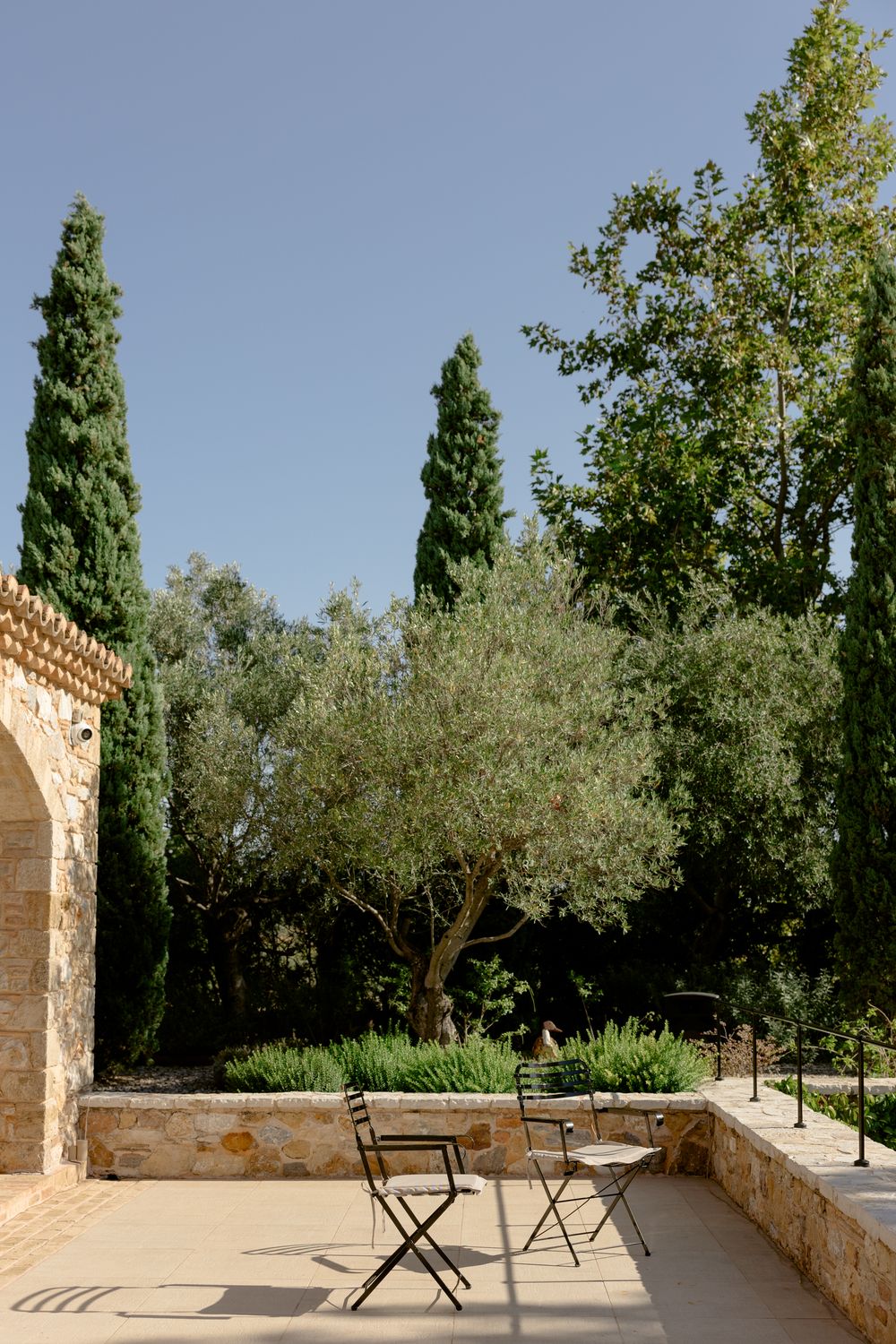 Mediterranean terrace with stone walls, cypress trees and outdoor seating under blue sky. Athens wedding photographer