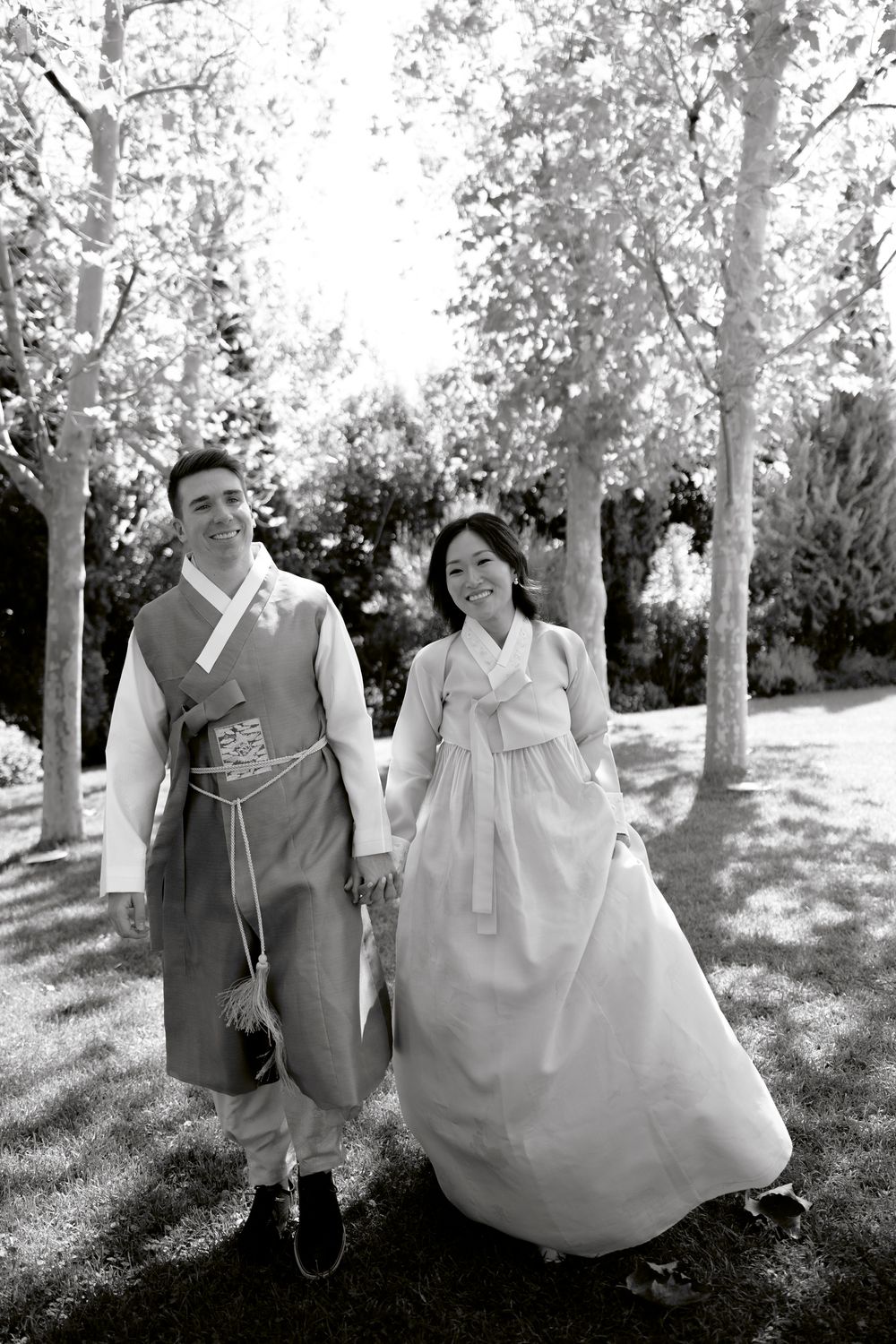 A couple in traditional Korean hanbok attire walking together in a sunlit garden path surrounded by trees. Athens wedding photographer
