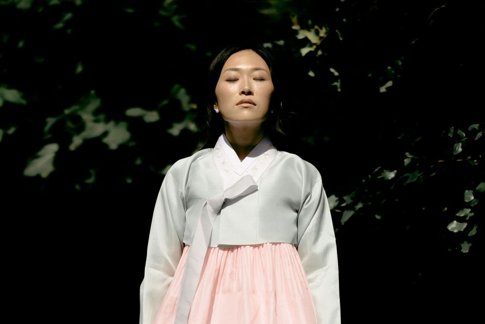 A person wearing a white and pink hanbok poses serenely in front of dark foliage. Athens wedding photographer