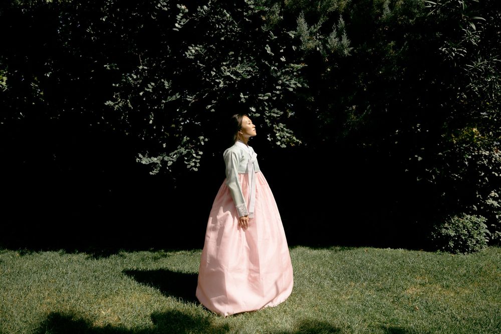 A figure in a pink traditional Korean hanbok dress stands alone in a garden setting. Athens wedding photographer