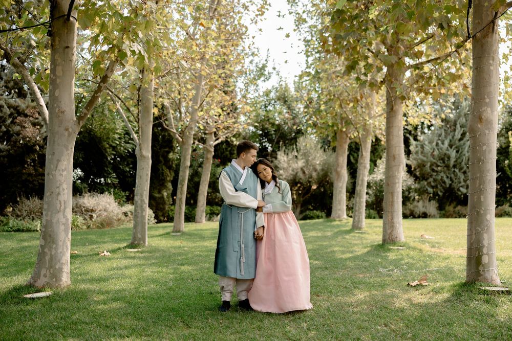 A couple in traditional Korean hanbok embrace in a tree-lined garden path. Athens wedding photographer