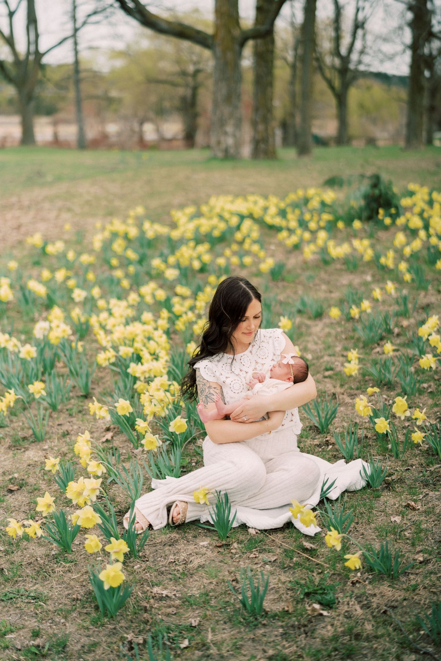 Spring nature scene with person in white dress sitting among blooming yellow daffodils in a wooded park setting.
