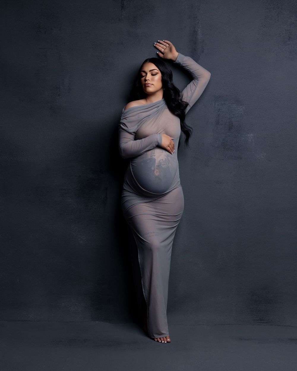 A maternity photo of someone wearing a sheer form-fitting gray dress posing elegantly against a dark background.