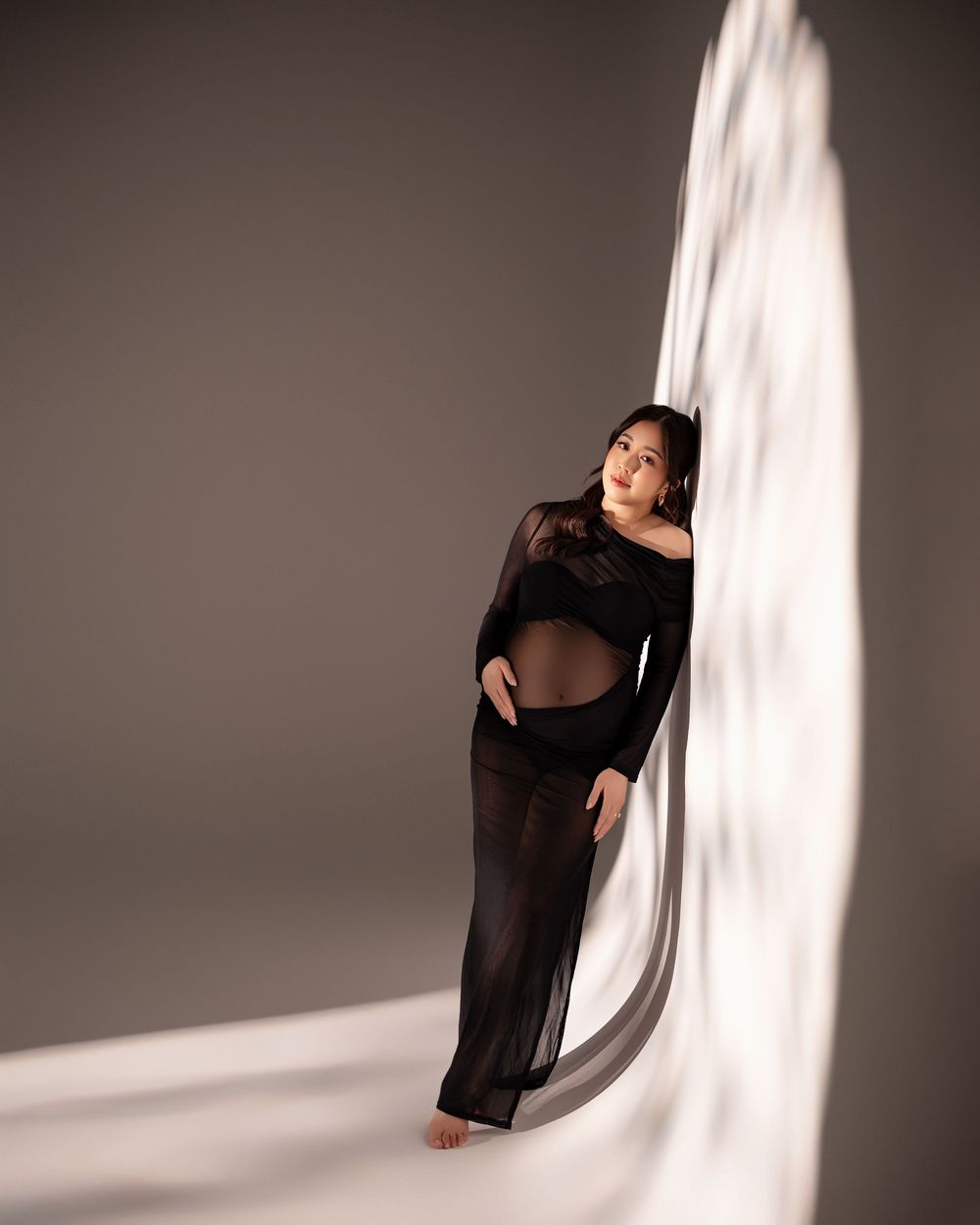 Model in sheer black gown poses elegantly against curved white backdrop in dramatic studio lighting.