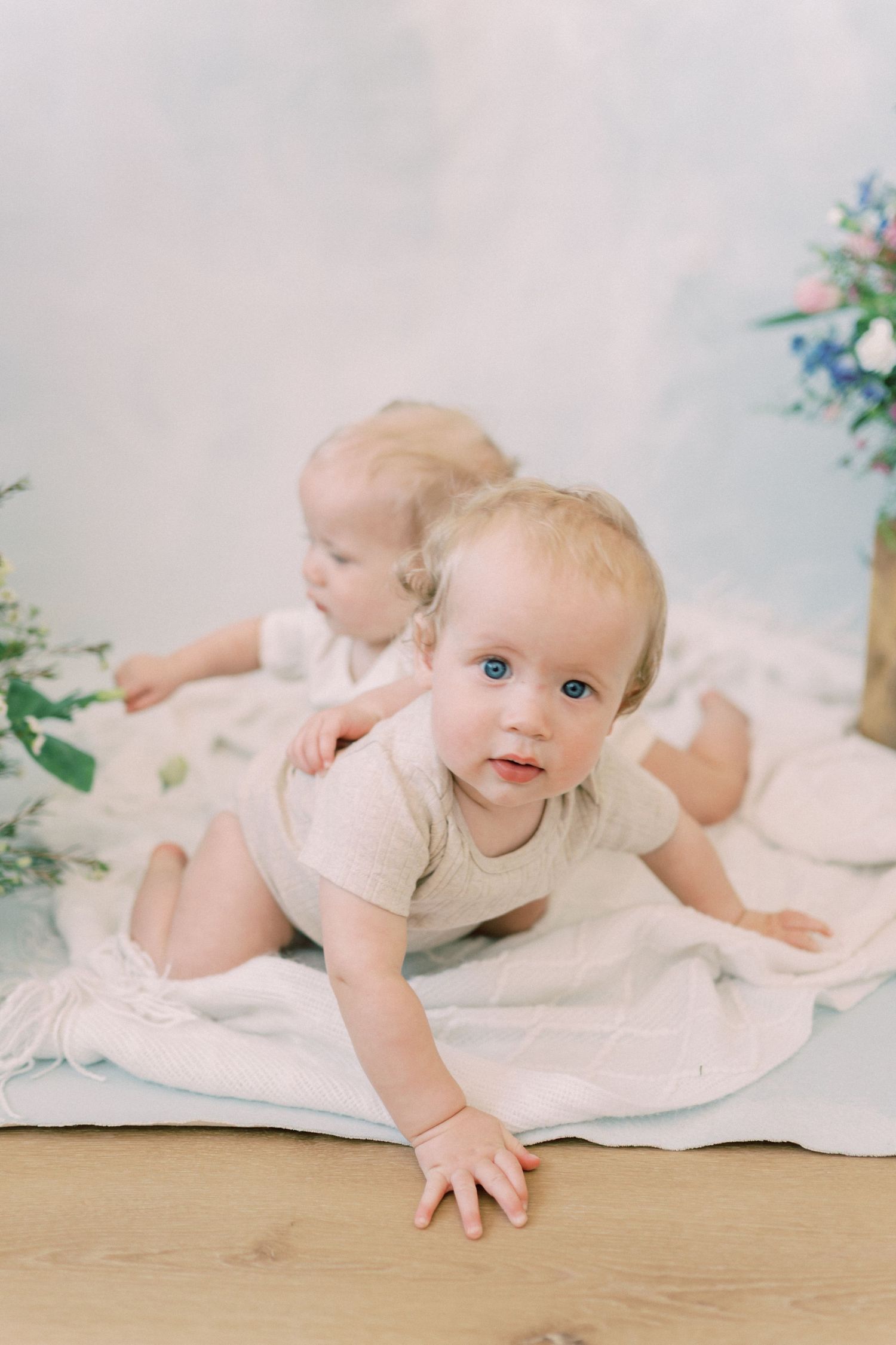 Two babies in light-colored outfits crawl on a white blanket near green plants against a neutral background.