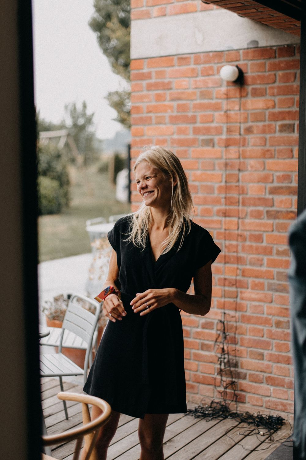 Woman in black dress laughing on outdoor deck with brick wall background.