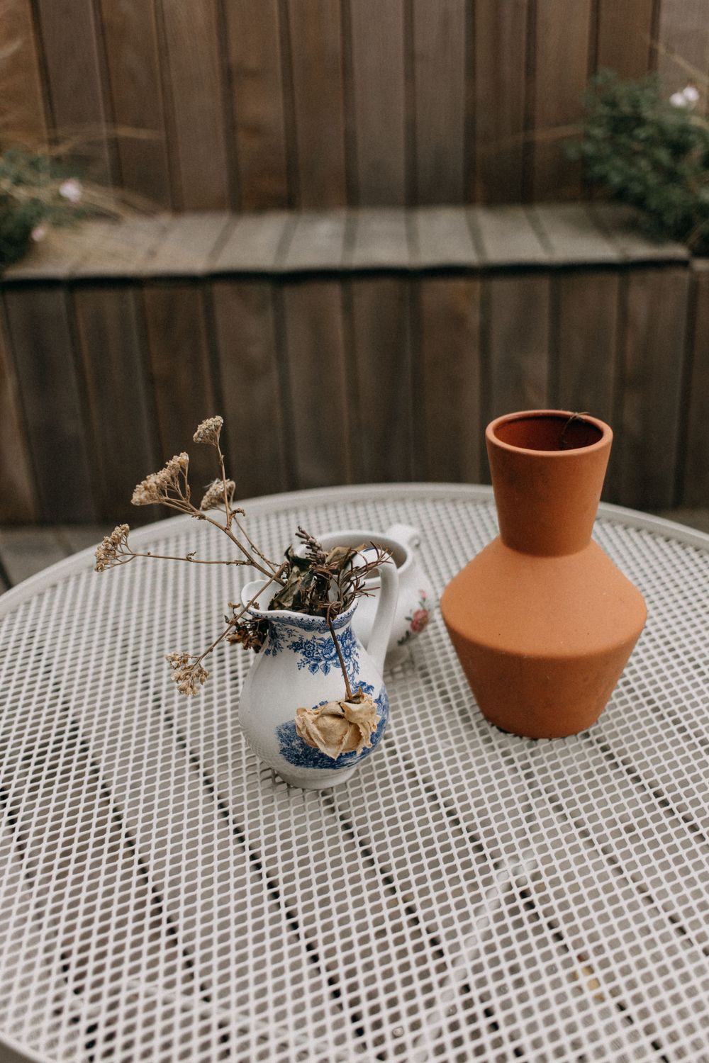 Terracotta vase and dried flowers on white mesh outdoor table.