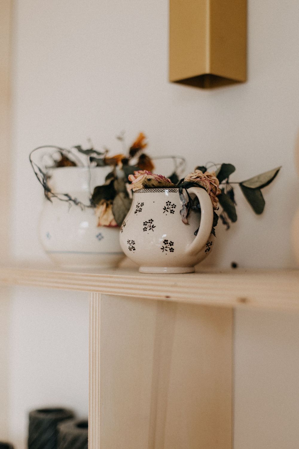 Two decorative teapots with floral designs sit on a wooden shelf against a white wall.