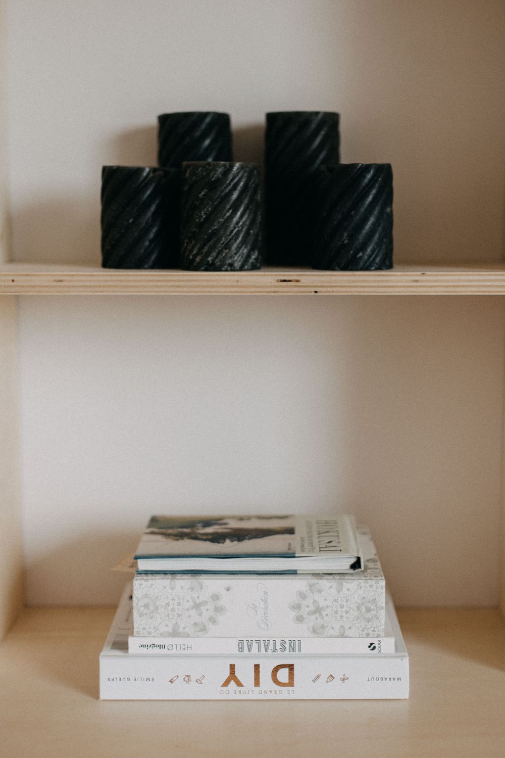 Black candles and stacked books arranged on minimal wooden shelving against neutral wall.