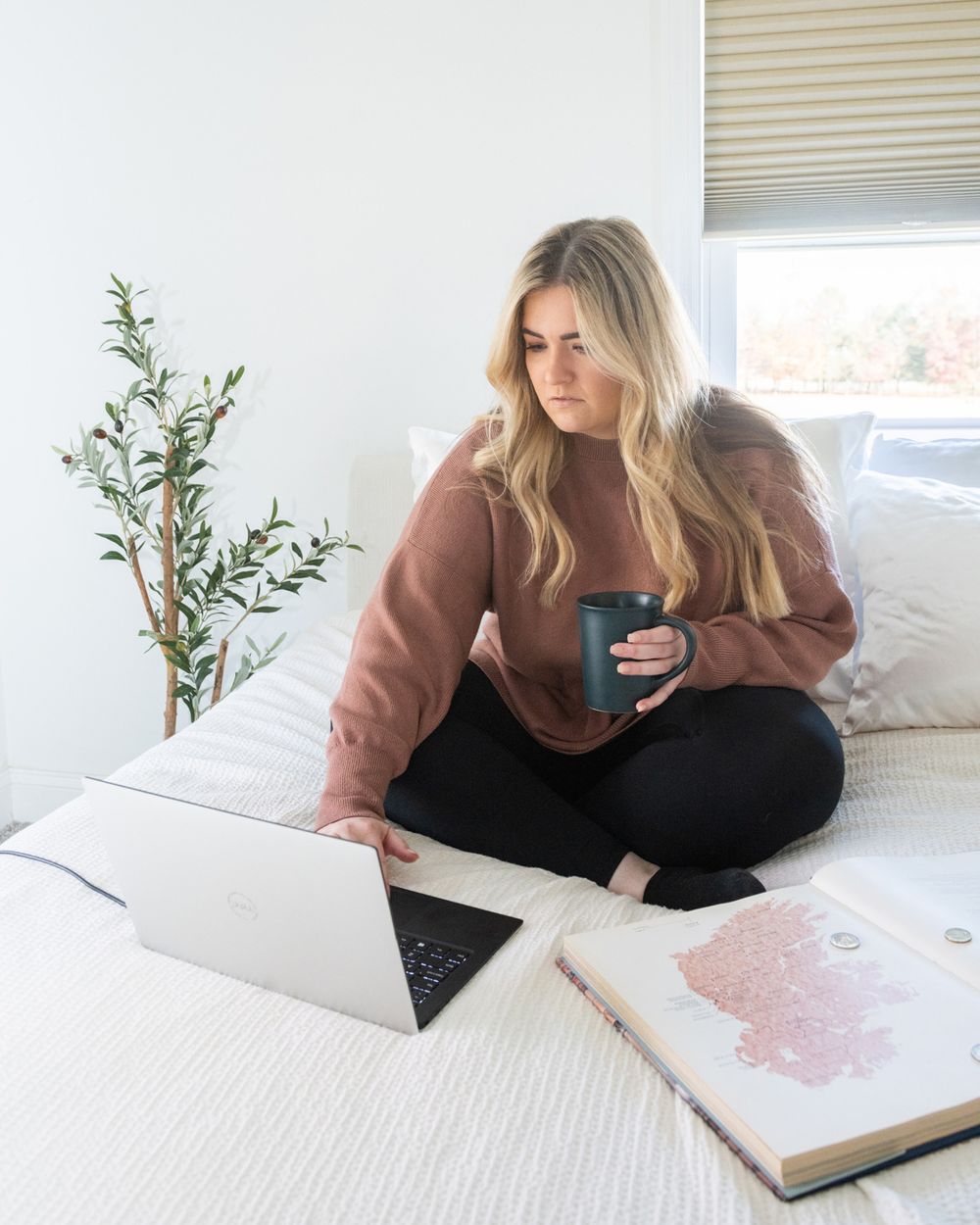 Person in brown sweater working on laptop in bright bedroom setting.