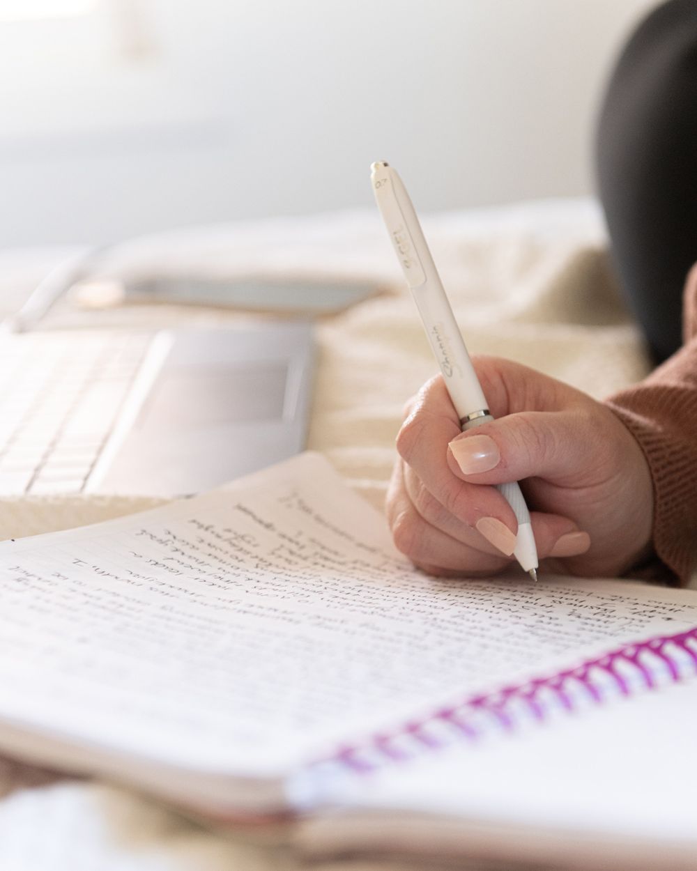 Hand holding white digital pen writing in spiral notebook with laptop nearby.