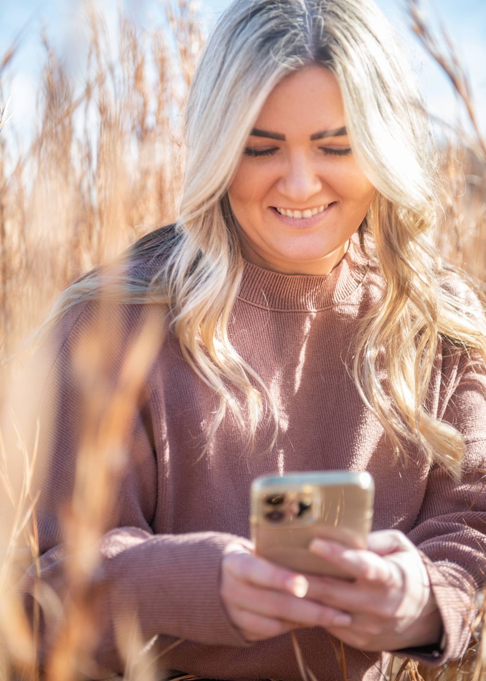 Woman in a mauve sweater smiling while looking at her phone amidst autumn wheat grass.