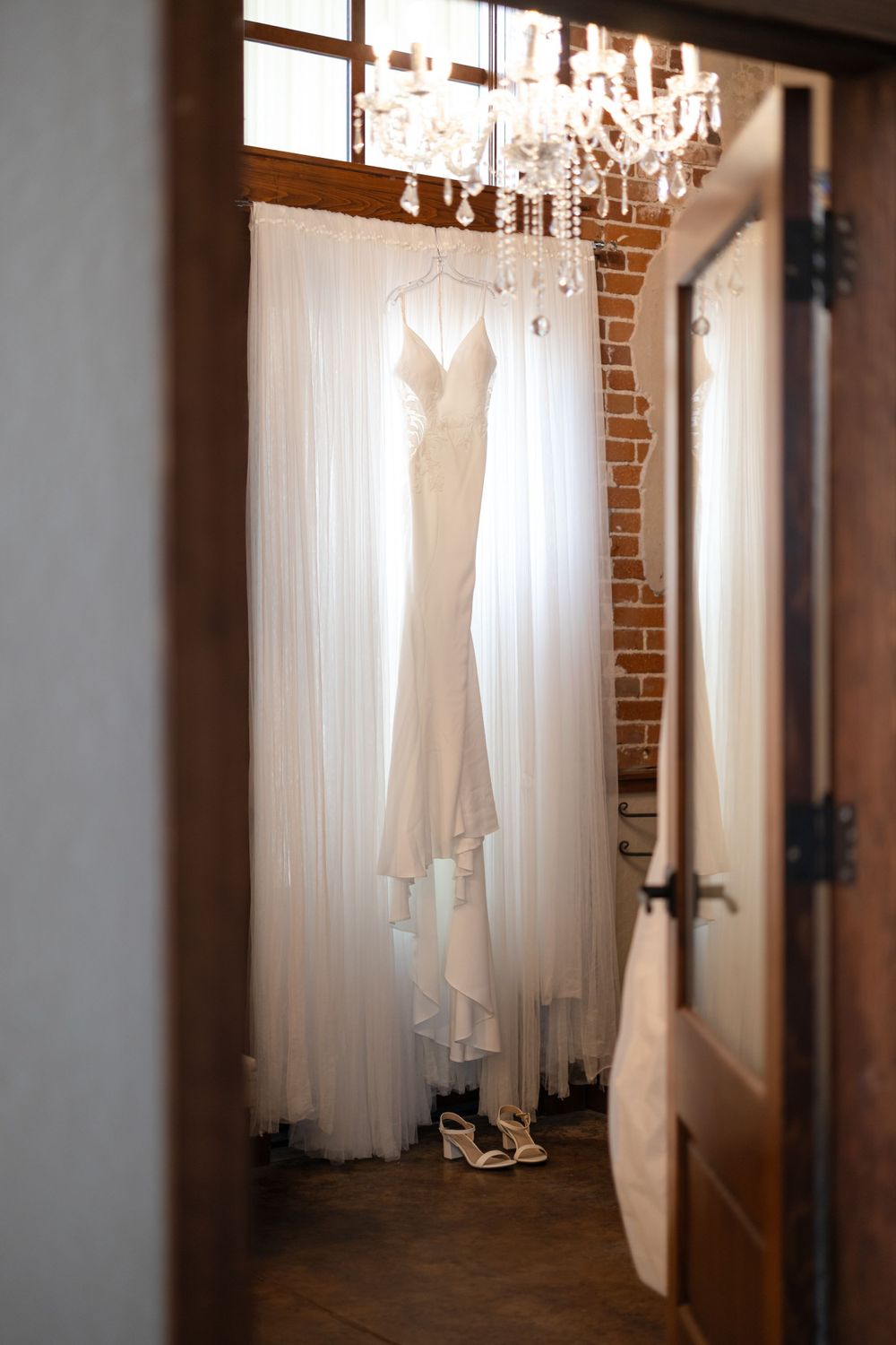 Ethereal wedding gown displayed in front of white curtain under crystal chandelier.