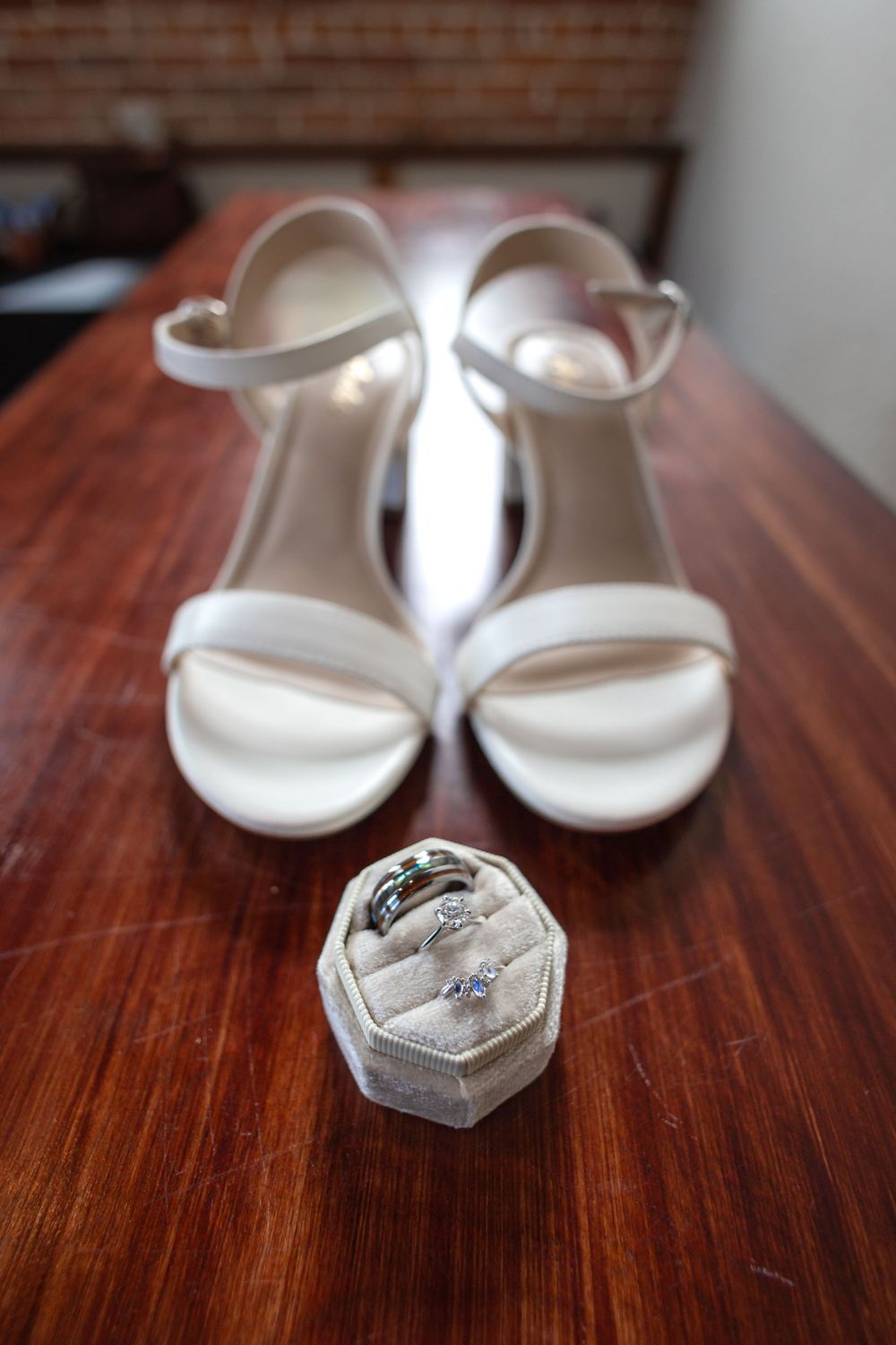 White bridal sandals and wedding rings displayed in hexagonal box on wooden surface.
