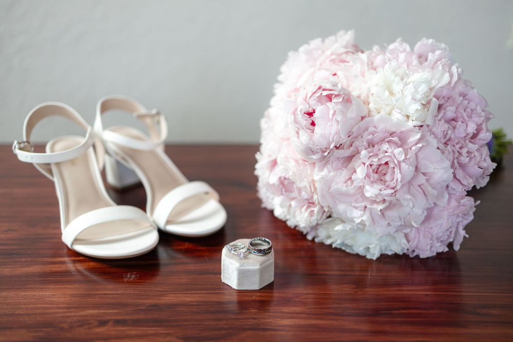 White wedding heels with pink peony bouquet and ring box on wooden table.