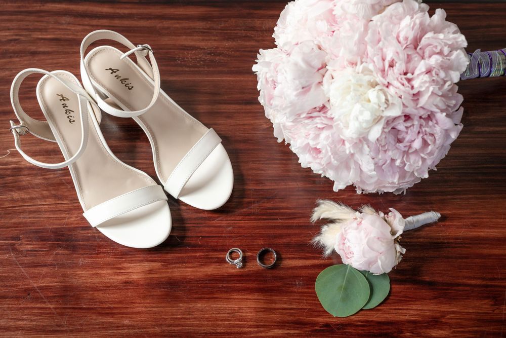 Bridal accessories including white sandals, rings, and pink peony bouquet arranged on wooden surface.