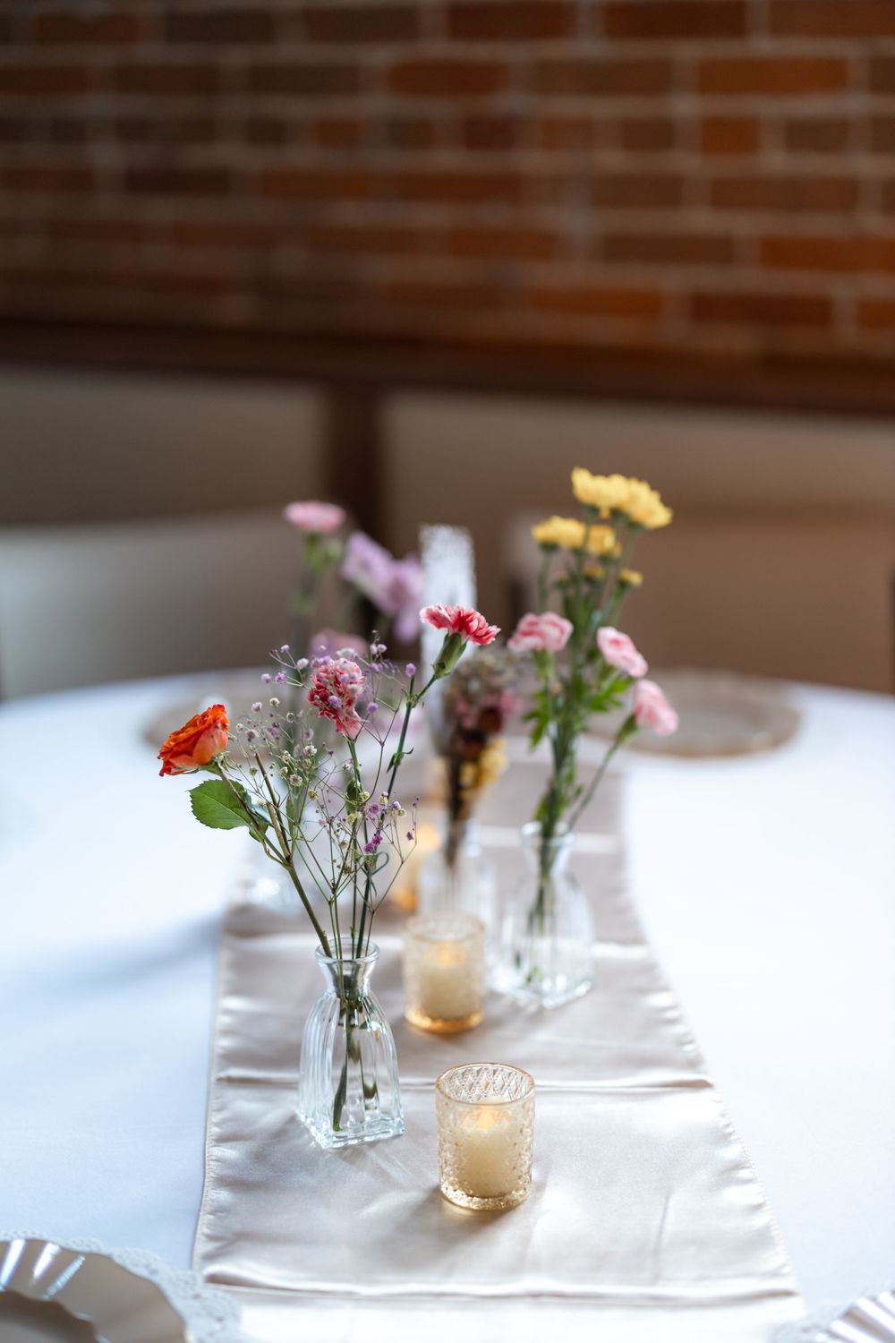 Small glass vases with wildflowers and tea lights arranged on white table runner.