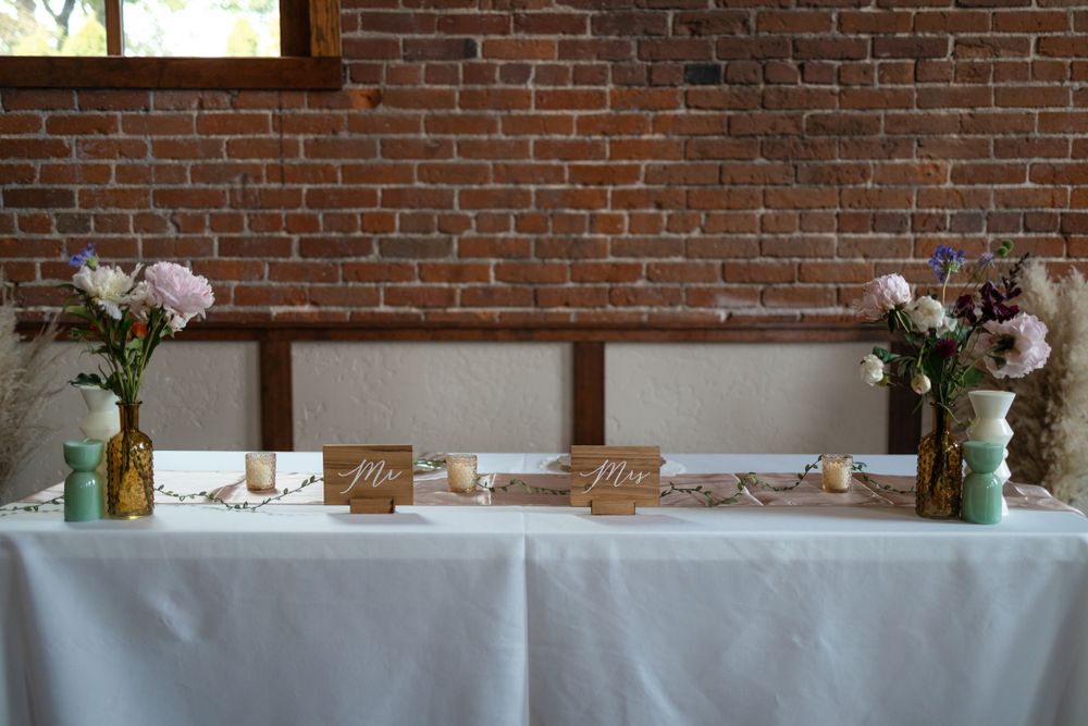 Reception table decorated with white linens, floral centerpieces, and decor against exposed brick wall.