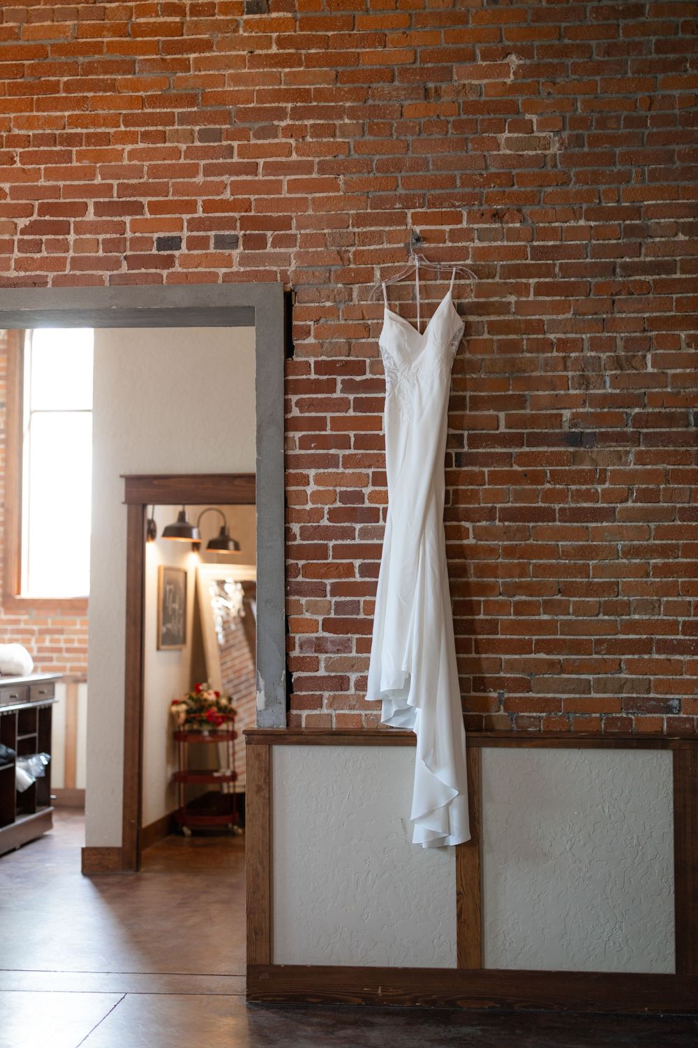 White wedding dress hangs elegantly against exposed brick wall in rustic venue interior.