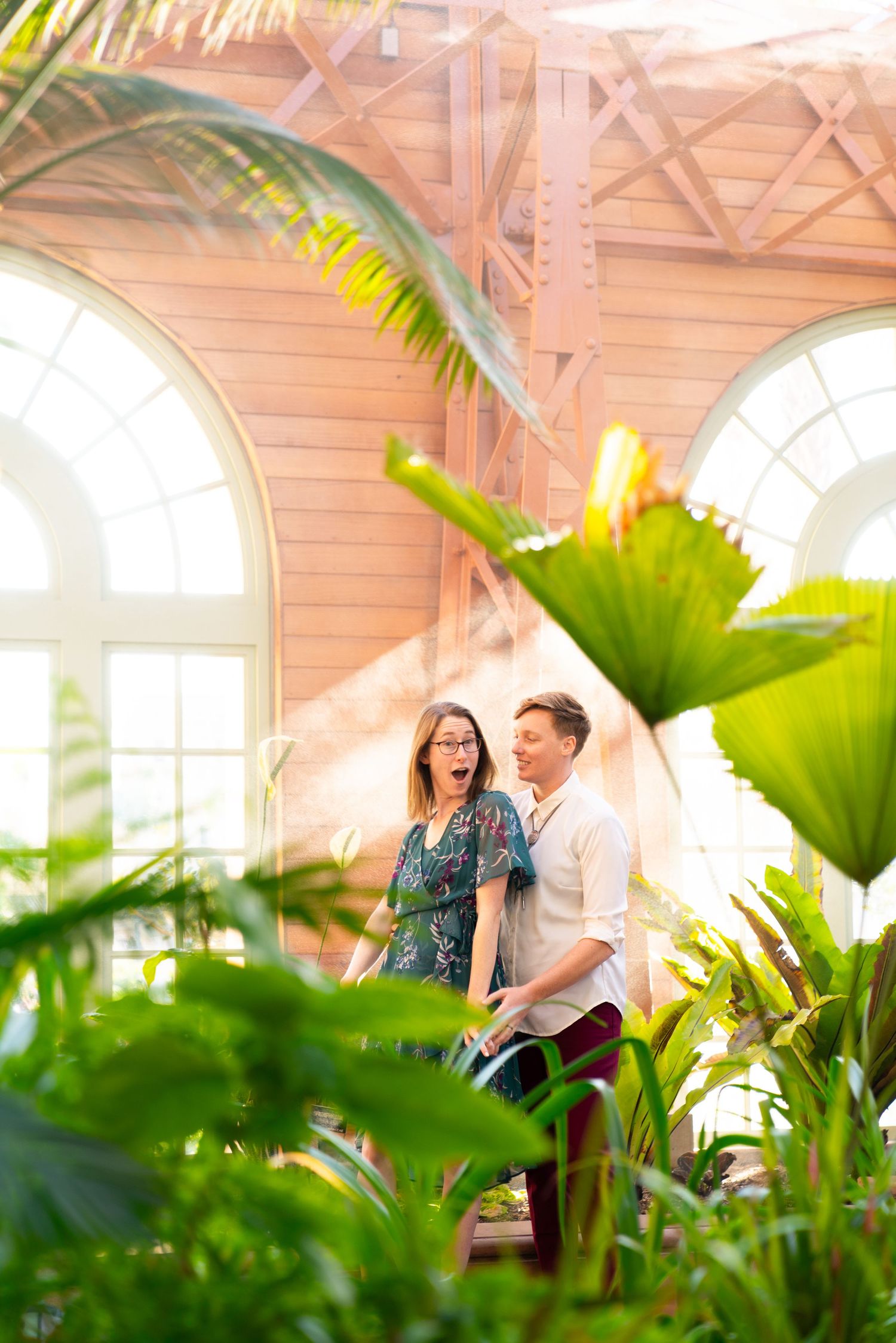 A couple shares a joyful moment surrounded by lush tropical plants in a sun-filled conservatory with arched windows.