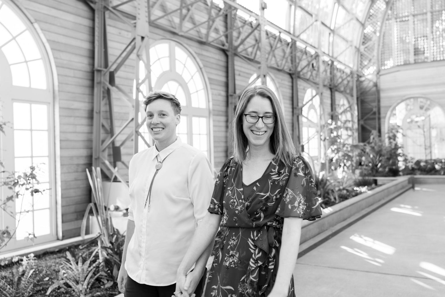 Two people stand smiling together in a bright greenhouse with arched windows and indoor plants.