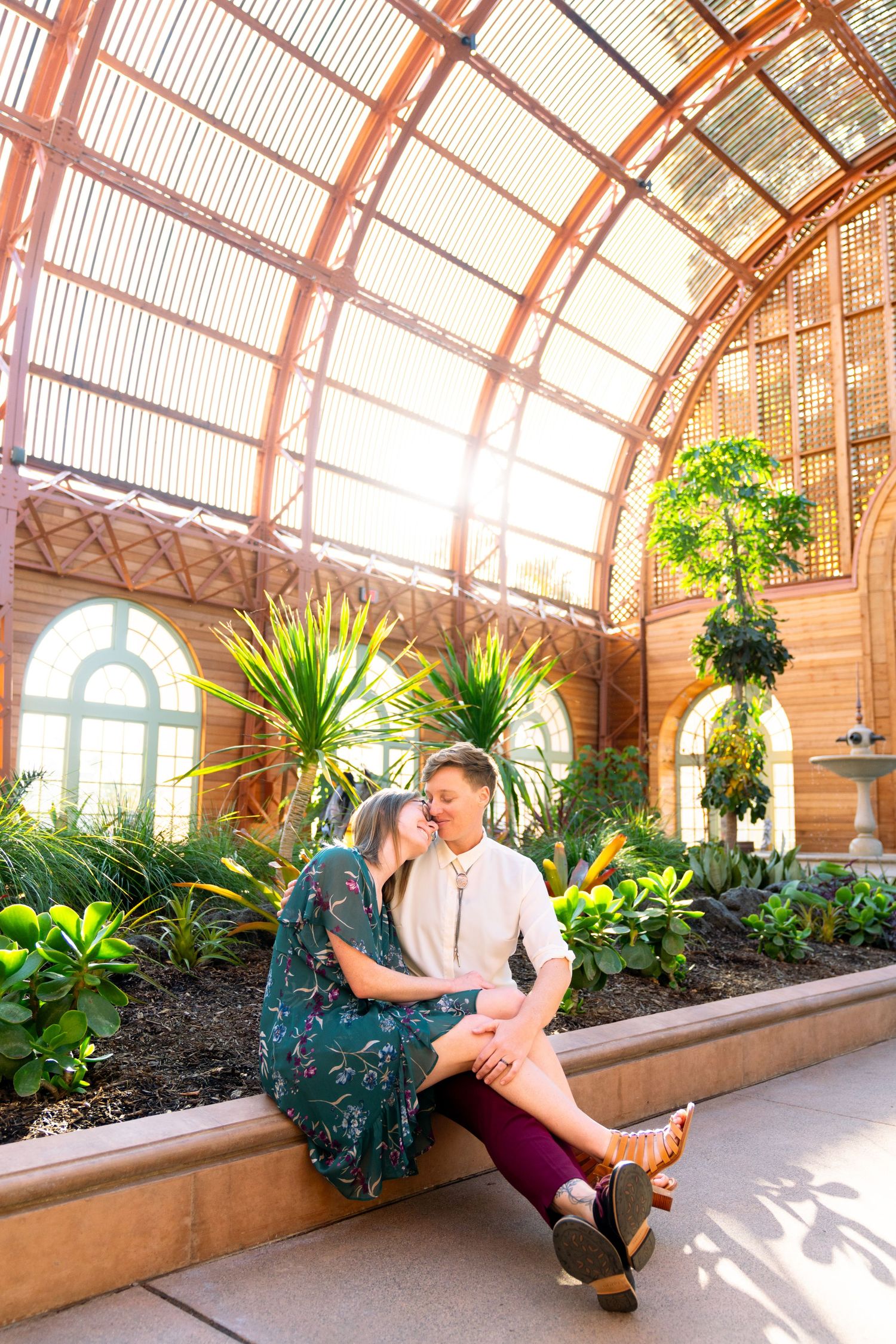 A couple sits together in a bright indoor garden conservatory with arched glass ceilings and tropical plants.