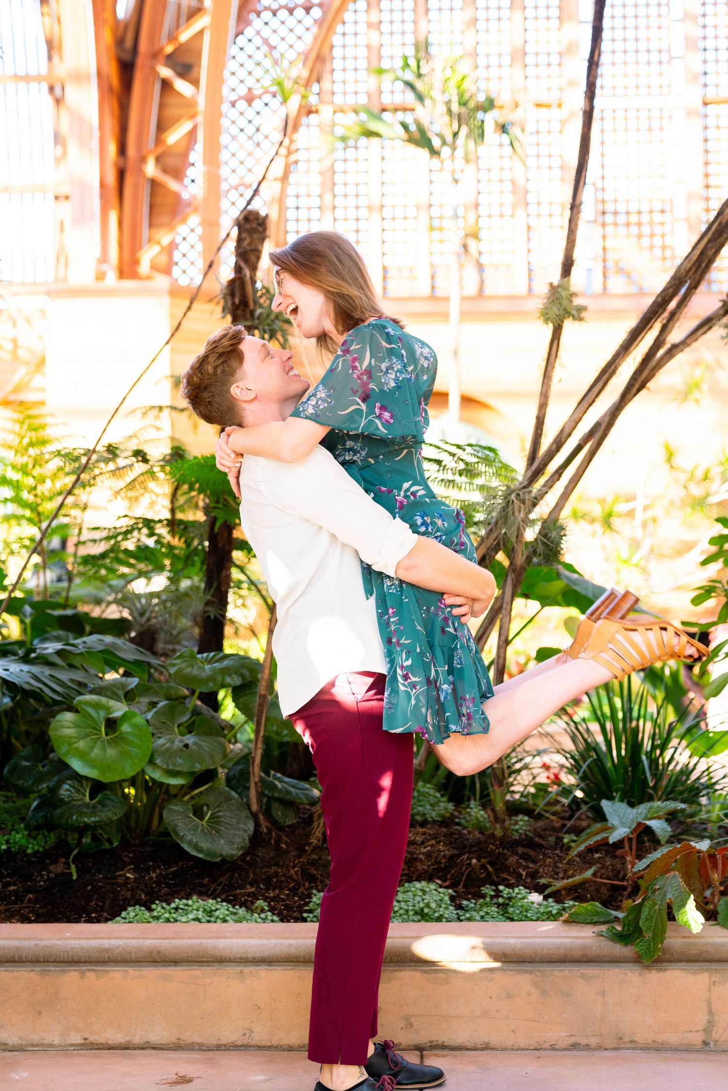 A couple embrace in a playful lift pose against a backdrop of tropical plants and lattice in an outdoor garden setting.