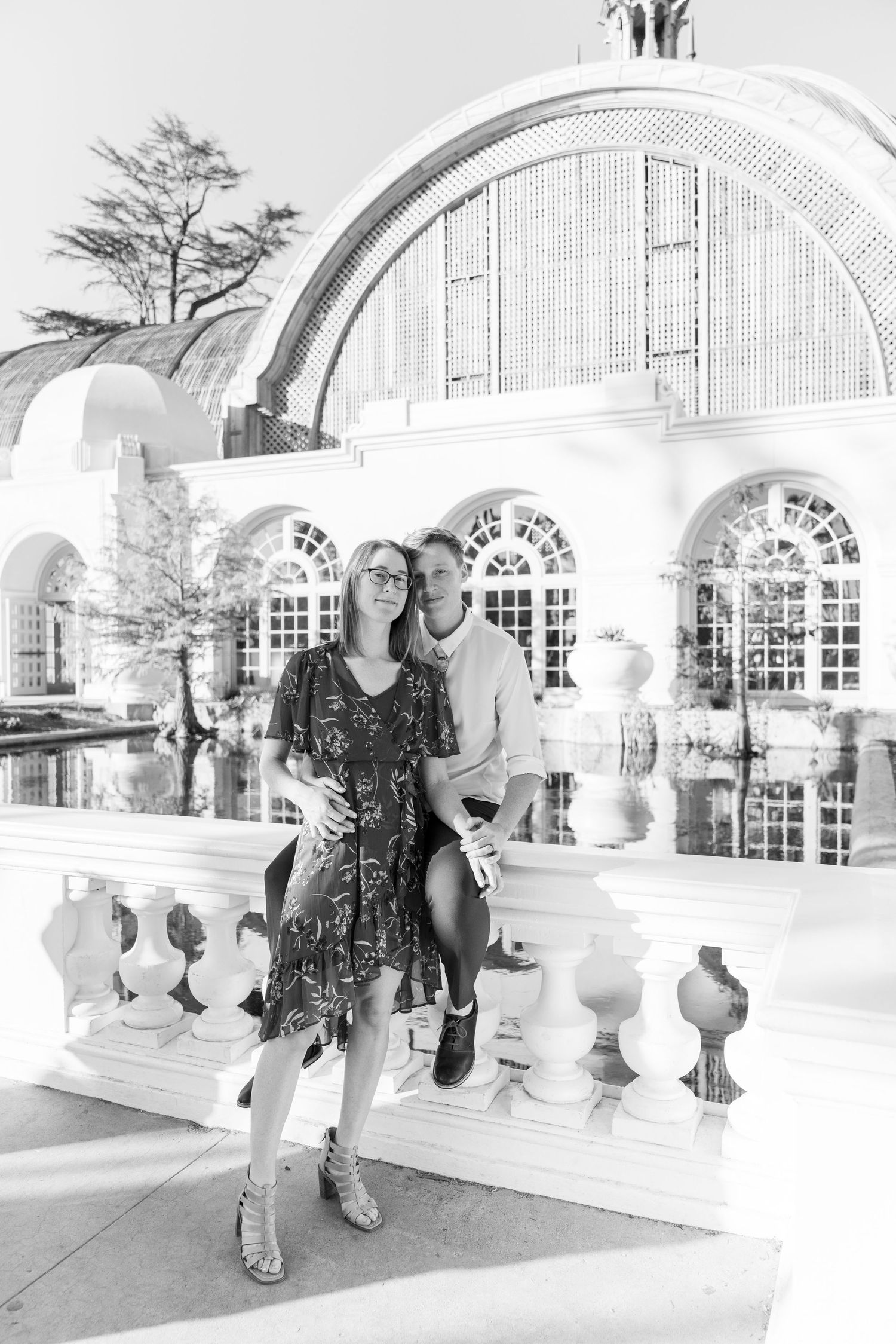 A couple poses together on a white stone balcony with elegant arched architecture and windows in black and white photography.