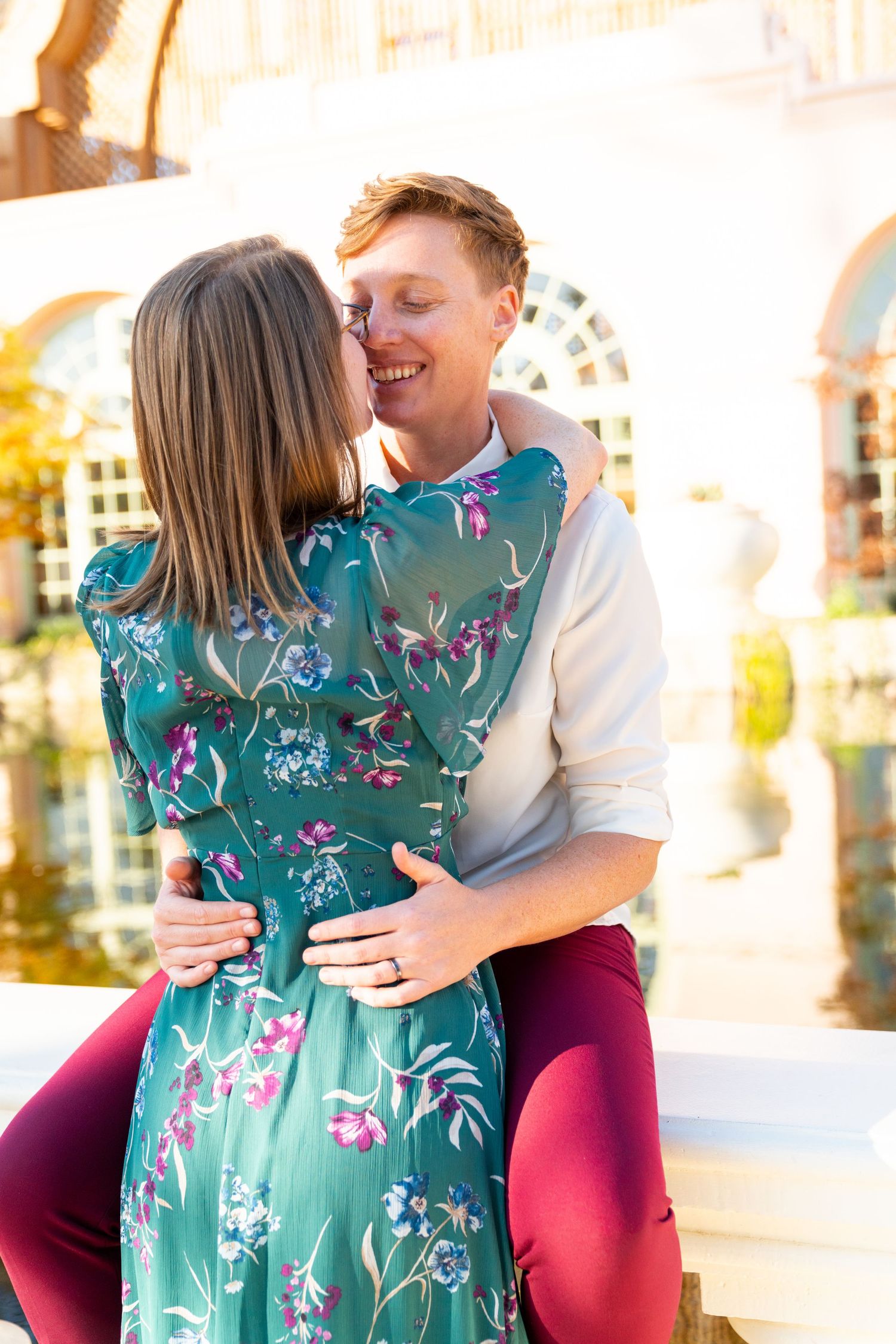 A couple embraces romantically in sunlight, with one wearing a teal floral dress and the other in a white shirt and burgundy pants.